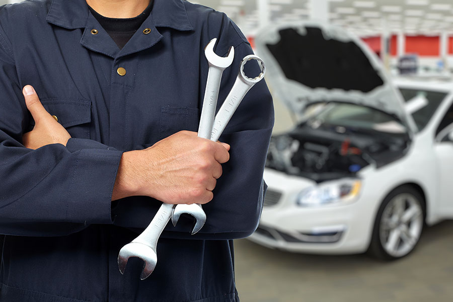 A mechanic’s arms are crossed, and his hands are holding several tools used to repair cars like the one in the background at a custom exhaust system shop in Decatur, IL.